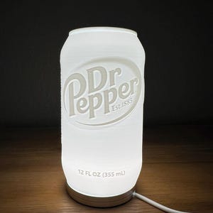 Può includere: Una lampada bianca illuminata a forma di lattina di soda Dr Pepper. La lampada presenta il logo Dr Pepper e la scritta "Est. 1885". La base della lampada riporta la scritta "12 FL OZ (355 ml)".