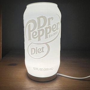 May include: A white, illuminated lamp shaped like a Dr Pepper Diet soda can. The lamp features the Dr Pepper logo and the words "Diet" and "12 FL OZ (355 mL)". The lamp is plugged in and sits on a wooden surface.