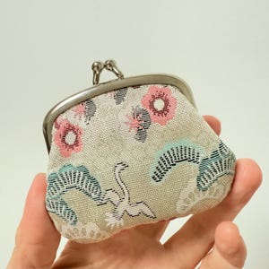Peut inclure: Un petit porte-monnaie beige avec un fermoir argenté. Le sac à main présente un motif floral avec des fleurs roses et blanches, et un motif de grue. Le sac est tenu dans une main.