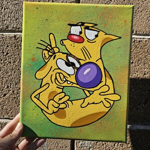 May include: A hand-painted canvas featuring a cartoon illustration of CatDog. The artwork depicts the two-headed character in yellow with a purple ball, set against a green background with orange paint splatters. The canvas is held against a brick wall.