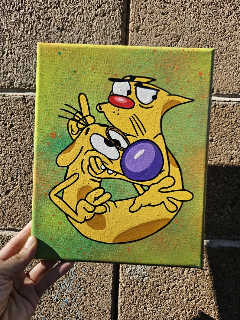 May include: A hand-painted canvas featuring a cartoon illustration of CatDog. The artwork depicts the two-headed character in yellow with a purple ball, set against a green background with orange paint splatters. The canvas is held against a brick wall.