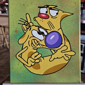 May include: A canvas painting featuring the cartoon characters Cat and Dog. The artwork depicts the characters in a playful embrace, with Cat's face close to Dog's. The background is a speckled green, and the characters are yellow with black outlines.