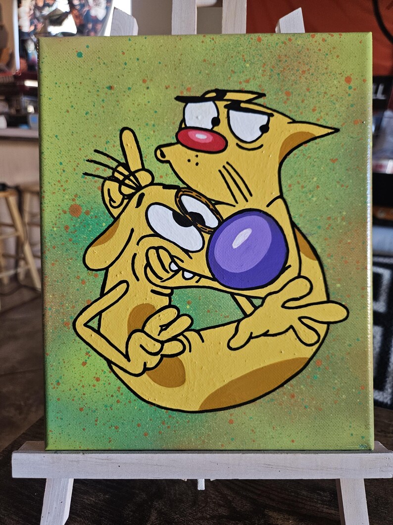 May include: A canvas painting featuring the cartoon characters Cat and Dog. The artwork depicts the characters in a playful embrace, with Cat's face close to Dog's. The background is a speckled green, and the characters are yellow with black outlines.
