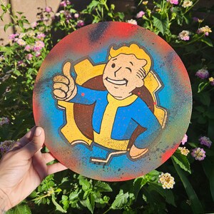 Puede incluir: Un disco redondo de madera pintado con un personaje de dibujos animados con un mono amarillo y una camisa azul, haciendo un pulgar hacia arriba. El personaje está rodeado de un engranaje amarillo con un fondo rojo, azul y amarillo.