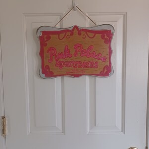 May include: A wooden sign with a white frame and pink lettering that reads "Pink Palace apartments".