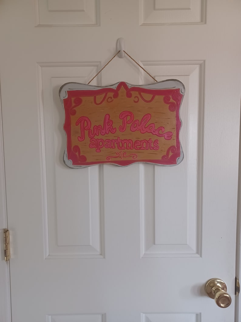 May include: A wooden sign with a white frame and pink lettering that reads "Pink Palace apartments".