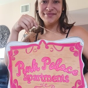 May include: A wooden sign with a pink frame and pink lettering that reads "Pink Palace Apartments".