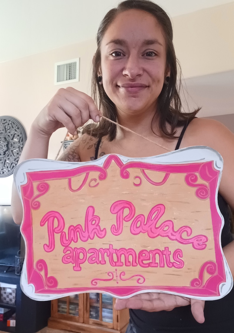 May include: A wooden sign with a pink frame and pink lettering that reads "Pink Palace Apartments".