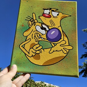 May include: A canvas painting featuring the cartoon characters Cat and Dog. The artwork depicts the characters in a yellow and black color scheme, set against a green background with paint splatters. The characters are embracing, with Dog holding up two fingers.