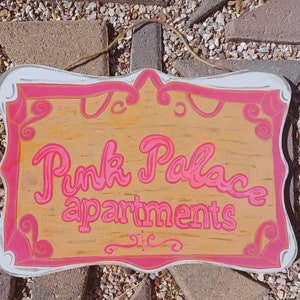 May include: A pink and white sign with the text "Pink Palace Apartments" in pink cursive lettering. The sign has a decorative border with swirls and a wood grain background.