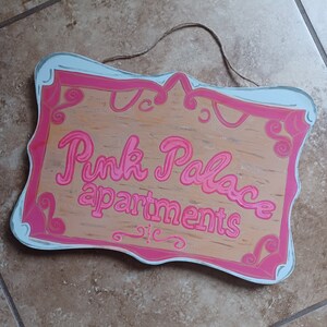 May include: A wooden sign with a pink and white painted frame. The sign says "Pink Palace apartments" in pink lettering.