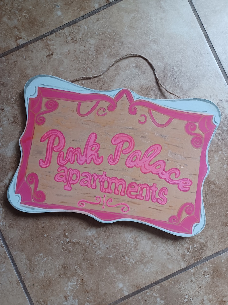 May include: A wooden sign with a pink and white painted frame. The sign says "Pink Palace apartments" in pink lettering.