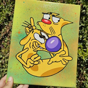 May include: A hand-painted canvas featuring the cartoon characters Cat and Dog. The artwork depicts the characters in a playful embrace, with Cat giving a peace sign. The background is a gradient of green and yellow with colorful paint splatters.