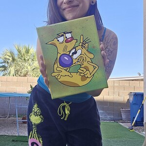 May include: A canvas painting featuring the cartoon characters Cat and Dog. The artwork depicts the duo in a playful pose against a green and yellow background. The painting is held up in front of a person.