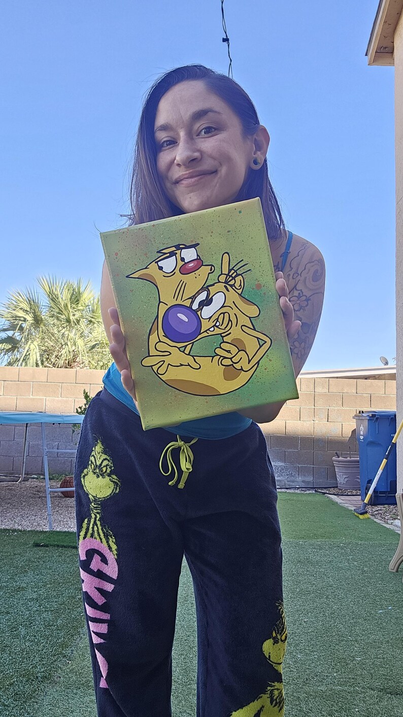 May include: A canvas painting featuring the cartoon characters Cat and Dog. The artwork depicts the duo in a playful pose against a green and yellow background. The painting is held up in front of a person.