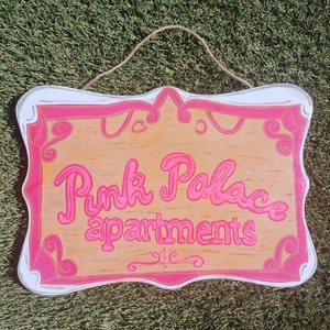 May include: Pink Palace Apartments sign with a white background and pink lettering. The sign is shaped like a heart and has a decorative border.