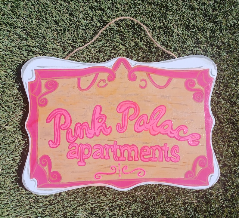 May include: Pink Palace Apartments sign with a white background and pink lettering. The sign is shaped like a heart and has a decorative border.