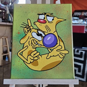 May include: A canvas painting featuring a cartoon image of CatDog, a yellow creature with two heads, one cat and one dog. The background is green with orange and brown paint splatters. The painting is displayed on a small wooden easel.