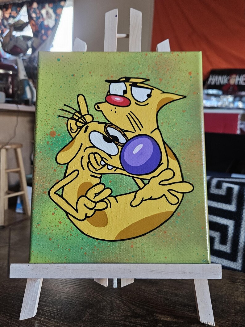 May include: A canvas painting featuring a cartoon image of CatDog, a yellow creature with two heads, one cat and one dog. The background is green with orange and brown paint splatters. The painting is displayed on a small wooden easel.
