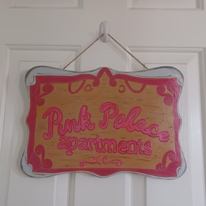 May include: A wooden sign with a pink and white painted frame and the words "Pink Palace apartments" in pink lettering.