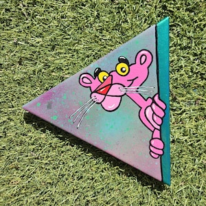 Puede incluir: Una obra de arte en lienzo triangular con el personaje de dibujos animados Pink Panther. La obra tiene un fondo degradado con colores rosa y turquesa. La Pantera Rosa es rosa con ojos amarillos y bigotes blancos.