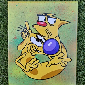 May include: A canvas painting featuring the cartoon characters Cat and Dog. The yellow characters are outlined in black, with Cat having a red nose and Dog with a purple nose. The background is a light green with colorful paint splatters.