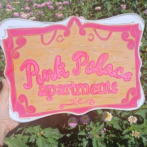 May include: A pink and white sign with the text "Pink Palace Apartments" in a whimsical font. The sign has a decorative border and is made of wood.