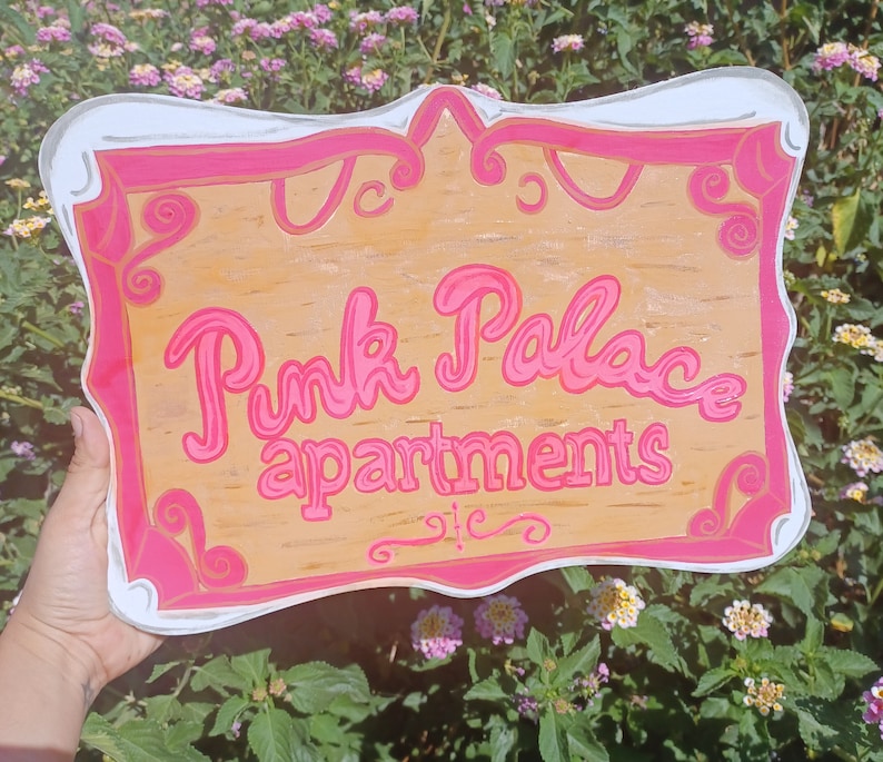 May include: A pink and white sign with the text "Pink Palace Apartments" in a whimsical font. The sign has a decorative border and is made of wood.