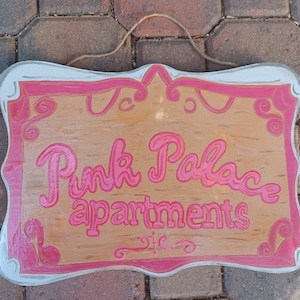 May include: A wooden sign with a pink painted border and the words "Pink Palace Apartments" in pink lettering.