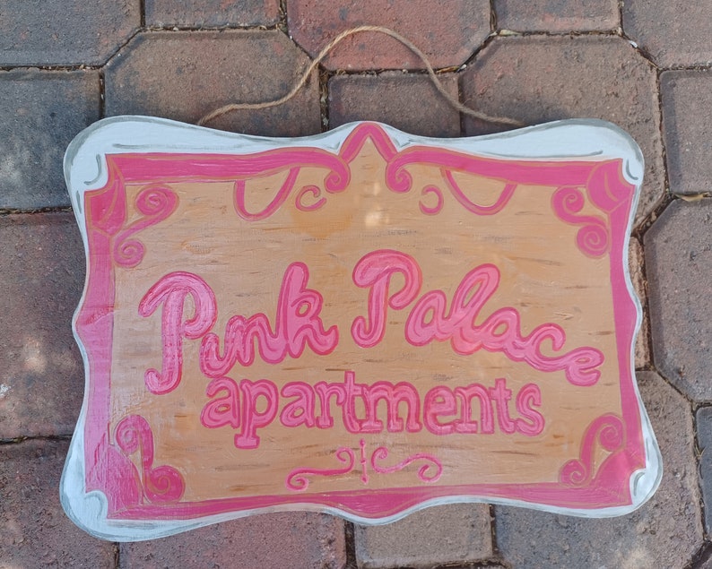 May include: A wooden sign with a pink painted border and the words "Pink Palace Apartments" in pink lettering.