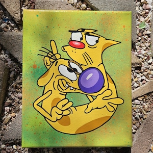 May include: A yellow cartoon painting of CatDog on a canvas. CatDog is a cartoon character with a cat and dog body. The background is a light green with orange and green paint splatters. The cat has a red nose and the dog has a purple nose.
