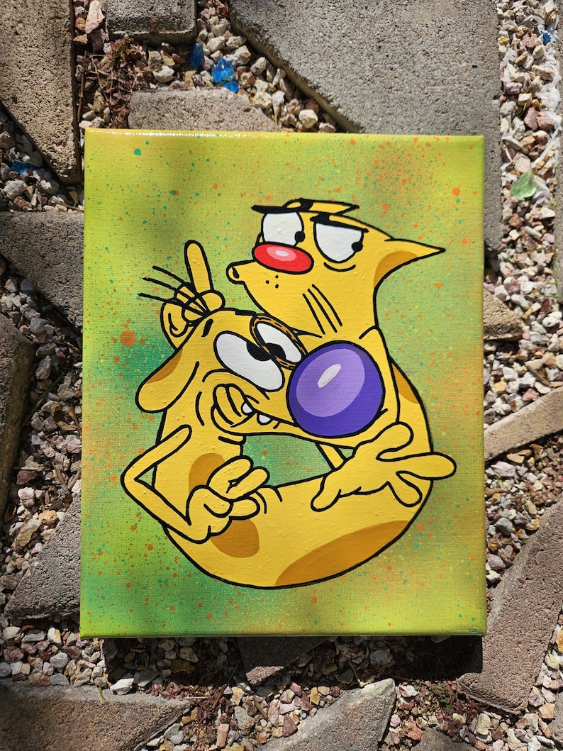 May include: A yellow cartoon painting of CatDog on a canvas. CatDog is a cartoon character with a cat and dog body. The background is a light green with orange and green paint splatters. The cat has a red nose and the dog has a purple nose.