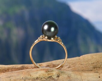 Puede incluir: Un anillo de oro con una gran perla verde oscuro engastada en el centro. El anillo está sobre un trozo de madera a la deriva con un fondo de montaña borroso.