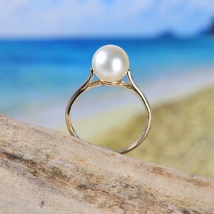 May include: A gold ring with a large, white pearl set in the center. The ring is resting on a piece of driftwood with a beach and ocean in the background.