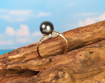 Puede incluir: Un anillo de oro con una gran perla negra engastada en el centro. El anillo está apoyado en un trozo de madera flotante.