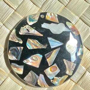 May include: A round, black pendant with a hole at the top. The pendant is made of iridescent abalone shell pieces that are set in a black resin.