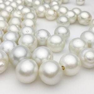 May include: A collection of lustrous, off-white pearls scattered on a white surface. The pearls vary slightly in size and shape, with a smooth, reflective surface. The image showcases the natural beauty and iridescence of the pearls.