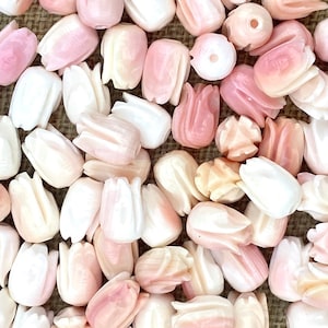 Puede incluir: Un conjunto de cuentas talladas en forma de tulipán en tonos blancos y rosas. Las cuentas, con un acabado pulido, están dispersas sobre una superficie texturizada. Las variadas tonalidades de rosa y blanco crean una estética suave y delicada. Cada cuenta mide aproximadamente 1,5 cm.