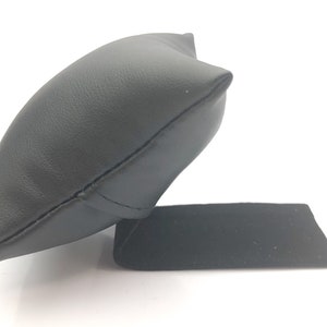 May include: Black leather watch pillow with a black velvet base. The pillow is shaped like a small cushion and is designed to hold a watch.