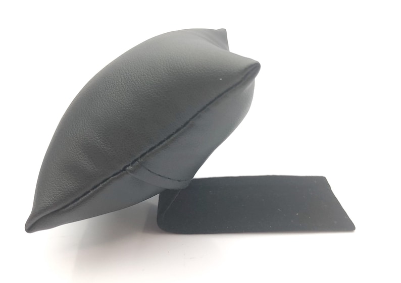 May include: Black leather watch pillow with a black velvet base. The pillow is shaped like a small cushion and is designed to hold a watch.