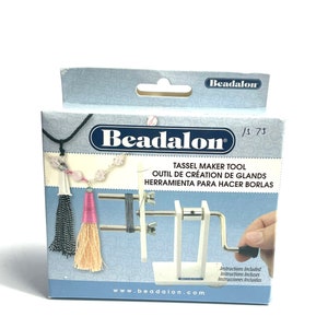 May include: A white plastic tassel maker tool with a metal crank. The tool is on a blue and white background with the text "Beadalon" and "Tassel Maker Tool" in black. The text "Outil de creation de glands" and "Herramienta para hacer borlas" is also visible in black.