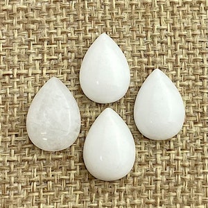 May include: Four white teardrop-shaped cabochons, likely made of quartz or agate, are arranged on a woven brown background.
