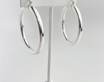 May include: A pair of polished silver hoop earrings displayed on a white jewelry stand. The earrings are round and have a smooth, reflective surface. The hoops are a medium size, suitable for everyday wear.