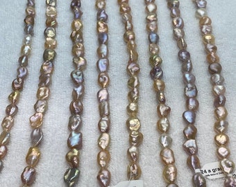 May include: Strands of Edison pearls in various colors, including peach, lavender, and cream, are displayed. The pearls have irregular shapes and a natural luster. Price tags indicate the cost per gram. The pearls are strung on thin, clear cords.