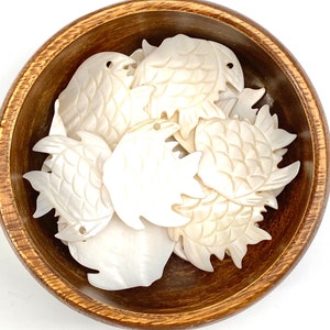 Puede incluir: Un bol de abalorios de nácar blanco con forma de pez. Los abalorios están tallados con un patrón de escamas detallado y tienen un agujero para encordar.