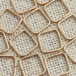 May include: A group of 10 gold-toned square jump rings. The rings are made of metal and have a smooth, polished finish. They are arranged in a random pattern on a beige fabric background.
