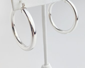May include: A pair of polished silver hoop earrings displayed on a white earring stand. The earrings are round with a thick, smooth design. The background is a plain white surface.