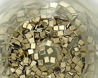 May include: A close-up view of a clear container filled with small, square, gold-colored mirror tiles.