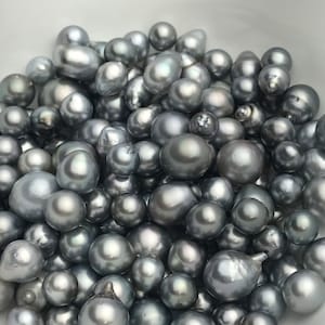 May include: A bowl filled with a variety of gray and silver iridescent pearls. The pearls are of different sizes and shapes, some are round, some are oval, and some are irregular.