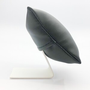 May include: A black leather watch pillow with a white base. The pillow is shaped like a rounded rectangle and is designed to hold a watch.